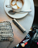 A Björk and Berries travel bottle placed on a table next to a coffee and a purse showing it is travel friendly.