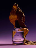 A tobacco leave leaning over a bottle of Björk and berries violet sky eau de perfume, with a purple background. 